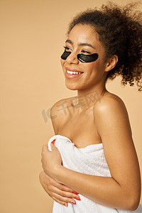 淋浴后裹着白毛巾的可爱的年轻女人微笑着站在一边，在米色背景下隔离的眼罩下涂上黑色