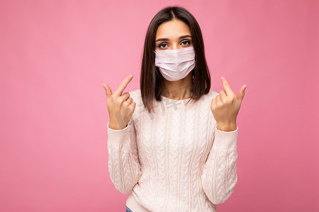 年轻漂亮的女人脸上戴着可重复使用的病毒防护面具，抵御粉红色背景墙上孤立的冠状病毒，并用手指指着自己