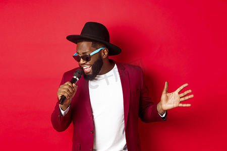 唱歌的男的摄影照片_热情的黑人男歌手在红色背景下表演，对着麦克风唱歌，穿着派对服装，站在红色背景上