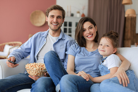 幸福的家庭，孩子坐在沙发上看电视吃爆米花，年轻的父母抱着女儿一起在沙发上放松。
