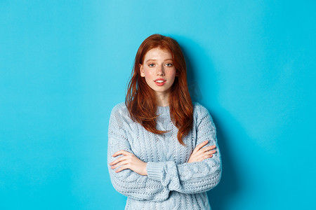 身穿温暖毛衣的红发少女站在蓝色背景上，自信地交叉双臂放在胸前