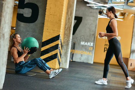 腹肌锻炼摄影照片_在私人教练的协助下，身穿运动服的年轻运动女性在健身房用健身球锻炼，进行腹肌锻炼