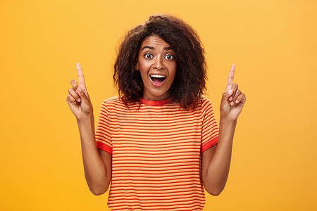 惊讶快乐热情可爱的非洲裔美国女性，头发卷曲，穿着条纹 T 恤，举起食指向上凝视，在橙色墙壁上看着相机，着迷和高兴
