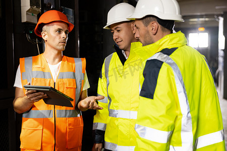 三名重工业工程师和工人站在管道制造厂，使用数字平板电脑进行讨论。