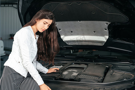 年轻女子站在车前，车库里打开引擎盖，看着引擎盖下
