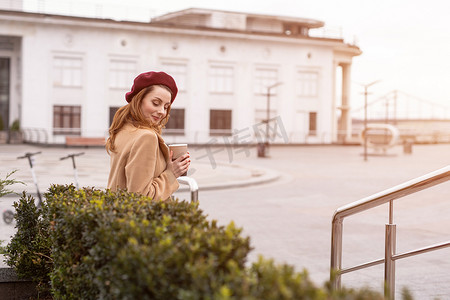 穿着秋装和米色的漂亮法式女人坐在大理石花坛上，在背景上分享或租用电动滑板车，等待她拿着咖啡杯的约会对象。