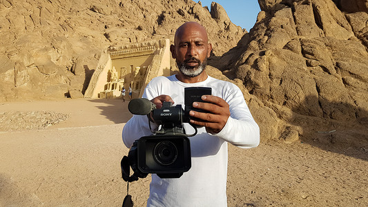 埃及，沙姆沙伊赫 - 2020 年 3 月 30 日。在沙姆沙伊赫的沙漠背景下，一名手里拿着索尼摄像机的成年埃及黑人摄像师正在杀害游客。