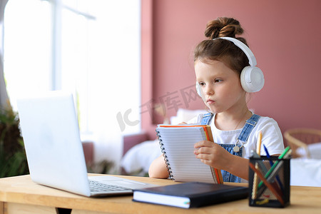 小孩子写字摄影照片_戴耳机的小女孩坐在办公桌前在笔记本上写字在线学习在家做运动，小孩子手写准备隔离作业，上网络课或在室内上课。