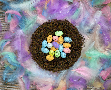 彩色羽毛摄影照片_鸟巢中色彩缤纷的复活节彩蛋，周围环绕着柔和的彩色羽毛和木制立方体，上面写着复活节快乐的顶视图，现代背景设计，复活节假期，春天，概念