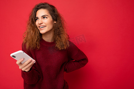 照片中，一头卷发的漂亮年轻女性身穿深红色毛衣，与红色背景隔离，手持手机，面带微笑