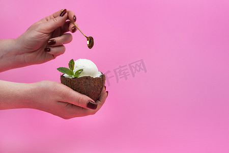 拿冰淇淋摄影照片_香草冰淇淋球放在新鲜椰子的一半里，手里拿着薄荷叶装饰，粉色背景上放着勺子