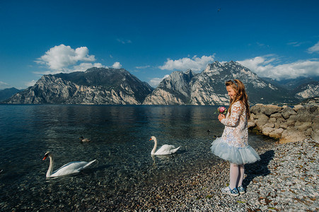一个穿着漂亮的白色连衣裙的小女孩沿着加尔达湖的堤岸散步。一个女孩在意大利的一座山和湖泊的背景下被拍照。Torbole