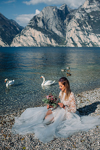 一个穿着漂亮的白色裙子的女孩坐在加尔达湖的堤岸上。一个女人在意大利的山湖背景下被拍到。Torbole