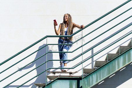 扎着彩色辫子的黑人女性，双脚搁在滑板上看着她的智能手机。