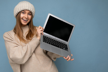 特写照片肖像美丽微笑的年轻金发女子在冬季温暖的针织米色帽子拿着电脑笔记本电脑看着相机穿着米色冬季毛衣隔离在浅蓝色墙壁背景在工作室