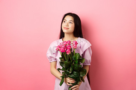 穿着可爱裙子的亚洲少女看着空荡荡的标志看起来很浪漫，拿着情人节鲜花礼物，从情人那里收到一束玫瑰，站在粉红色的背景上