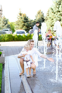 年轻的金发妈妈和小孩坐在喷泉附近。