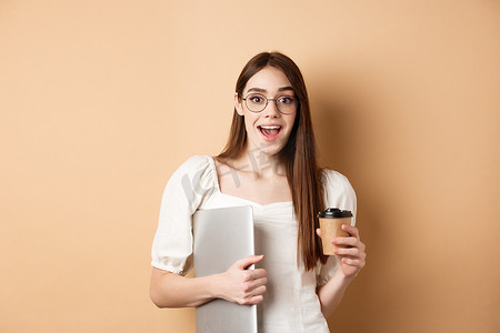 戴着眼镜的兴奋女人从外卖杯里喝咖啡，拿着笔记本电脑，开心地笑着，站在米色背景上