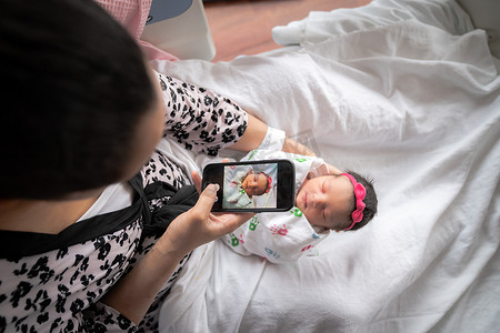一位穿着粉色动物印花浴袍的母亲坐在病床上，抱着她刚出生的女婴裹在毯子里，同时用手机拍照与家人和朋友分享。