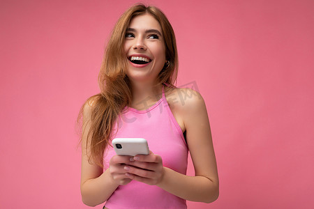 照片中，微笑、快乐、漂亮的年轻金发女子穿着粉红色上衣，在粉红色背景下与世隔绝，手里拿着空的空间，使用手机短信抬头做梦