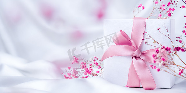 白色礼物盒，白色丝绸面料背景上有粉色丝带和小粉色花朵。