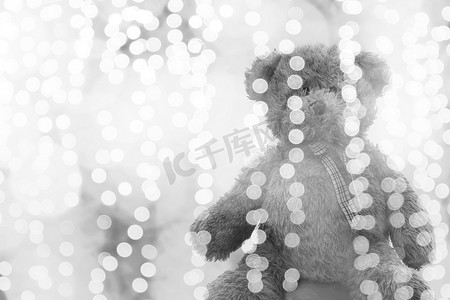 泰迪熊娃娃在灯光线散景银色明亮的圣诞节或新年快乐背景中，熊孤独地坐在闪闪发光的灰色白色背景中模糊的散景明亮（选择性聚焦）