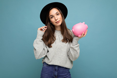 钱箱icon摄影照片_正面微笑的可爱年轻漂亮女人的照片，她留着深色头发，穿着休闲装，戴着黑色帽子，在蓝色背景上与复制空间隔开，拿着粉红色的小猪盒来赚钱。