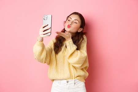 漂亮的轻浮女孩在智能手机上自拍，在视频聊天中发送飞吻，站在粉红色背景下