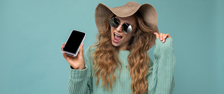 美丽快乐的年轻金发女子戴着太阳镜和夏季帽子的全景照片，在蓝色背景中被隔离，复制空间拿着智能手机，手拿手机，屏幕显示空白，张着嘴看着相机