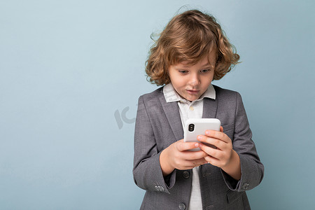 短卷发男孩摄影照片_照片中，一头卷发、身穿灰色西装、手持和使用蓝色背景中突显的手机的英俊惊奇男孩看着智能手机交流 vie 短信