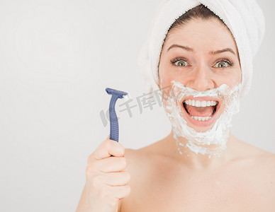 头戴毛巾、脸上剃须泡沫的快乐白人女性拿着白色背景上的剃刀