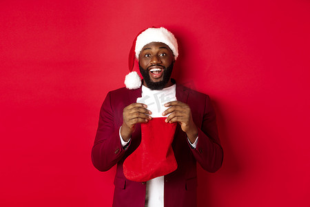 圣诞袜里有礼物摄影照片_兴奋的黑人打开圣诞袜，里面放着礼物和糖果，笑得很开心，站在红色背景下的圣诞帽里