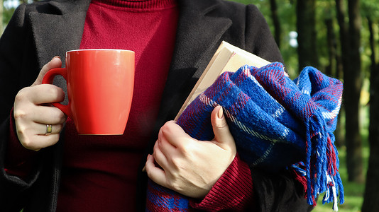 花纹红摄影照片_公园里一位穿着毛衣和黑色外套的女人手里拿着一杯红咖啡和一本书，书上有蓝色格子羊毛毯或格子。