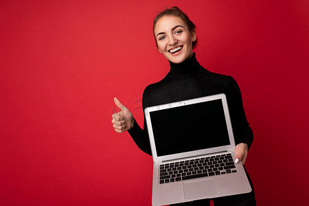 照片中，美丽微笑、快乐的黑发年轻女子手持电脑笔记本电脑，屏幕空空如也，身穿黑色长袖，看着相机，在红墙背景中竖起大拇指
