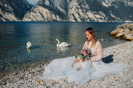 一个穿着漂亮的白色裙子的女孩坐在加尔达湖的堤岸上。一个女人在意大利的山湖背景下被拍到。Torbole