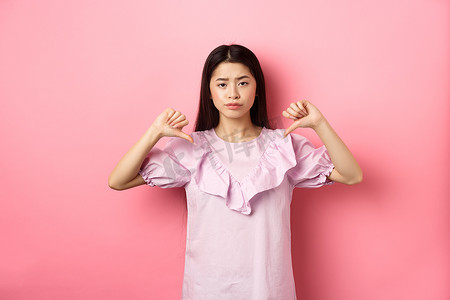 持怀疑态度和失望的亚洲女性站在粉色背景上，不情愿地笑，不喜欢产品，表现出消极情绪，大拇指朝下