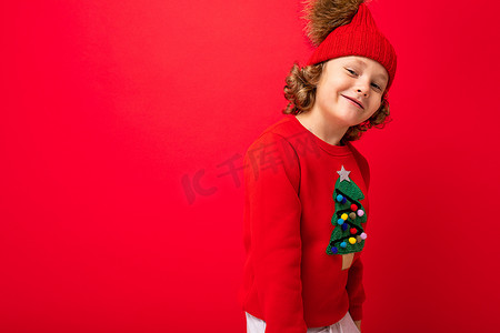 可爱的金发男孩，戴着温暖的帽子，穿着红色背景的圣诞毛衣，脸上挂着微笑