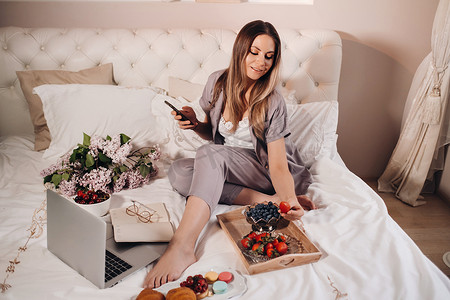 一个女孩晚上坐在床上，手里拿着智能手机吃着草莓，一个女孩在床上睡前吃糖果。