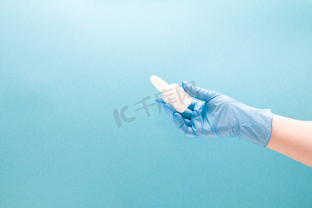 一只戴蓝色一次性医用手套的女手拿着一个白色塑料瓶，里面有滴鼻液，没有帽子，蓝色背景复制空间，滴鼻液，鼻药