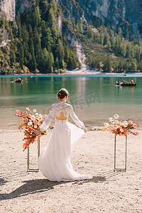 白色连衣裙摄影照片_在意大利的 Lago di Braies，美丽的新娘穿着带袖子和蕾丝的白色连衣裙，在拱门的背景上放着一束黄色的秋天花束参加仪式。