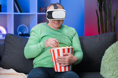 老人坐在沙发上拿着爆米花，他戴着虚拟现实眼镜，举起手来选择 VR 屏幕。