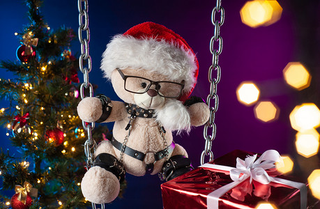 圣诞老人帽子里的泰迪熊是圣诞树背景下 BDSM 游戏的圣诞礼物
