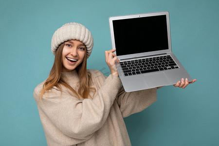 美丽迷人迷人漂亮微笑快乐年轻深色金发女学生冬季温暖针织米色帽子拿着电脑笔记本电脑看着相机穿着米色冬季毛衣隔离在工作室的浅蓝色背景