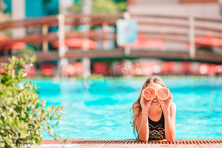 背景游泳池中，小女孩用靠近眼睛的橙色半边遮住眼睛