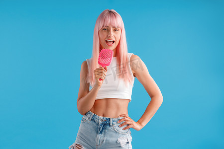 粉色丝滑丝滑摄影照片_站在蓝色工作室背景中，一头粉红色头发的快乐女人拿着梳子，在里面唱歌