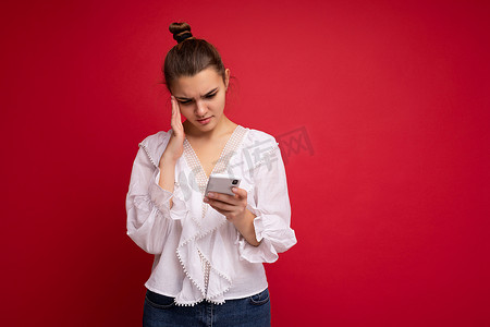 思念的摄影照片_身穿白色衬衫的漂亮年轻、心烦意乱的黑发女人站在红色背景上，通过手机看手机屏幕，阅读互联网上的新闻