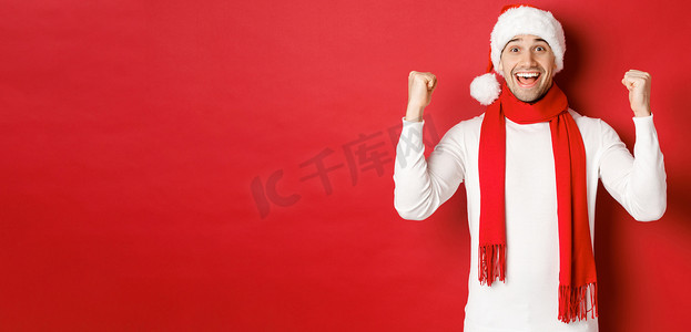 戴着圣诞帽和围巾的快乐而兴奋的男人的画像，欢欣鼓舞并赢得了一些东西，庆祝新年，站在红色背景上