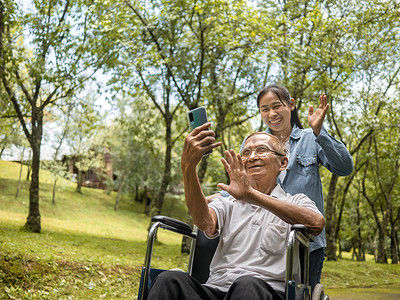 坐在轮椅上的祖父和孙女在公园里通过智能手机与亲戚进行视频通话。