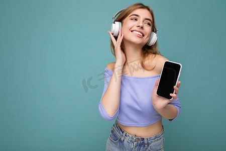 照片中，美丽、快乐、微笑的年轻女子穿着时尚的休闲服，隔离在背景墙外，手持并使用手机，戴着白色蓝牙耳机听音乐，并有趣地看着旁边