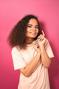 美丽的黑皮肤非洲裔美国年轻女孩穿着粉红色背景中突显的桃红色 T 恤，轻轻地看着镜头。